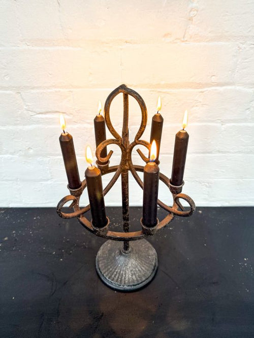 A five-arm wrought iron candelabra with a distressed dark brown finish, holding five dark brown taper candles that are lit, set on a dark surface against a white brick wall.