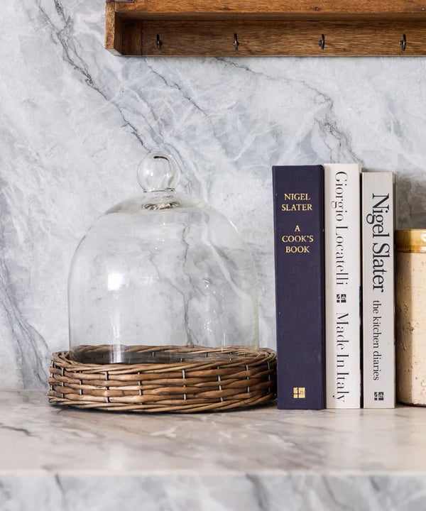 Glass cloche on a marble surface with cookbooks stacked behind it. Riviera Cloche Dome on Base Small by French Country Collections 